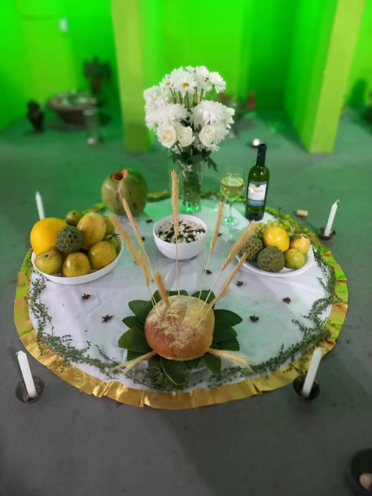 Oferenda no templo sobre toalha branca com babado dourado, com flores brancas, pão, espigas de trigo, frutas amarelas, vinho branco e velas