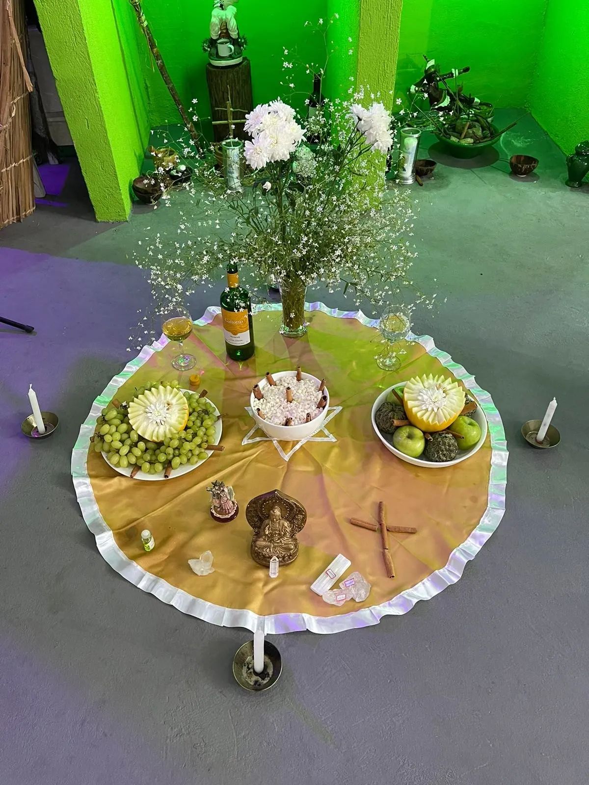 Oferenda no templo sobre toalha dourada com babado branco, com flores brancas, uvas, melões, vinho, velas e uma pequena cruz