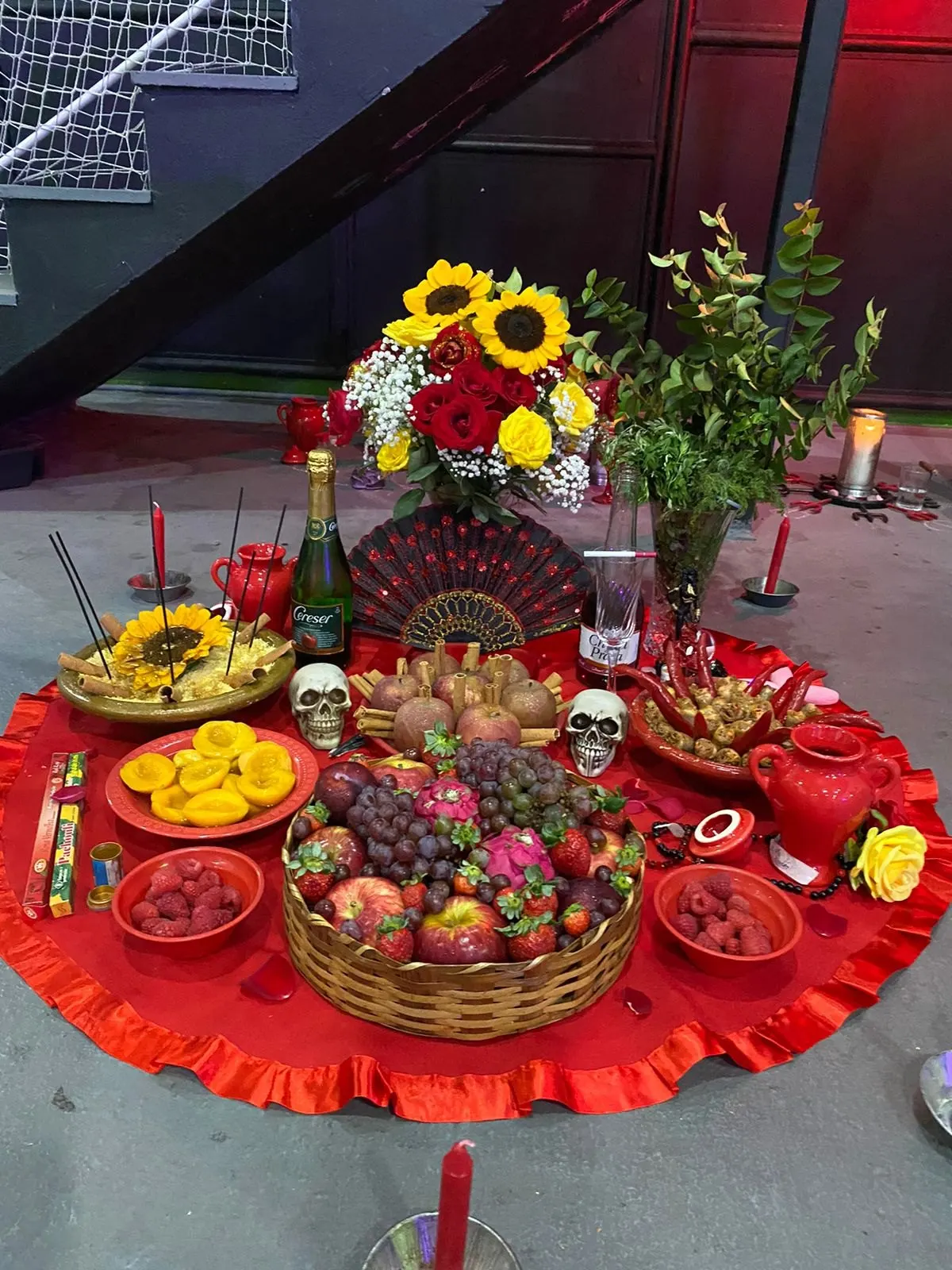 Oferenda a Pombagira no templo, sobre toalha vermelha, com girassóis e rosas, leque, duas caveiras, cesta de frutas, pêssegos, morangos e espumante