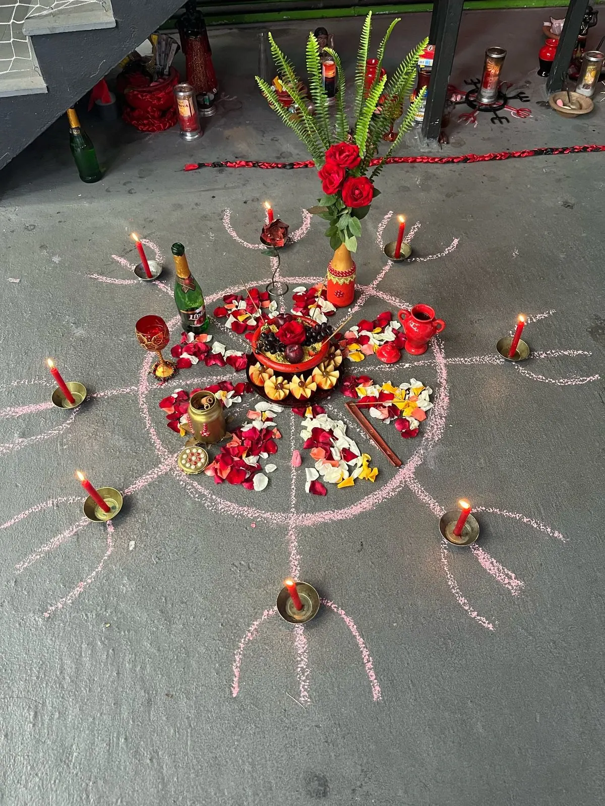 Oferenda a Pombagira no chão do templo, cercada de linhas riscadas a giz com velas vermelhas nas pontas, rosas vermelhas, espumante e pétalas