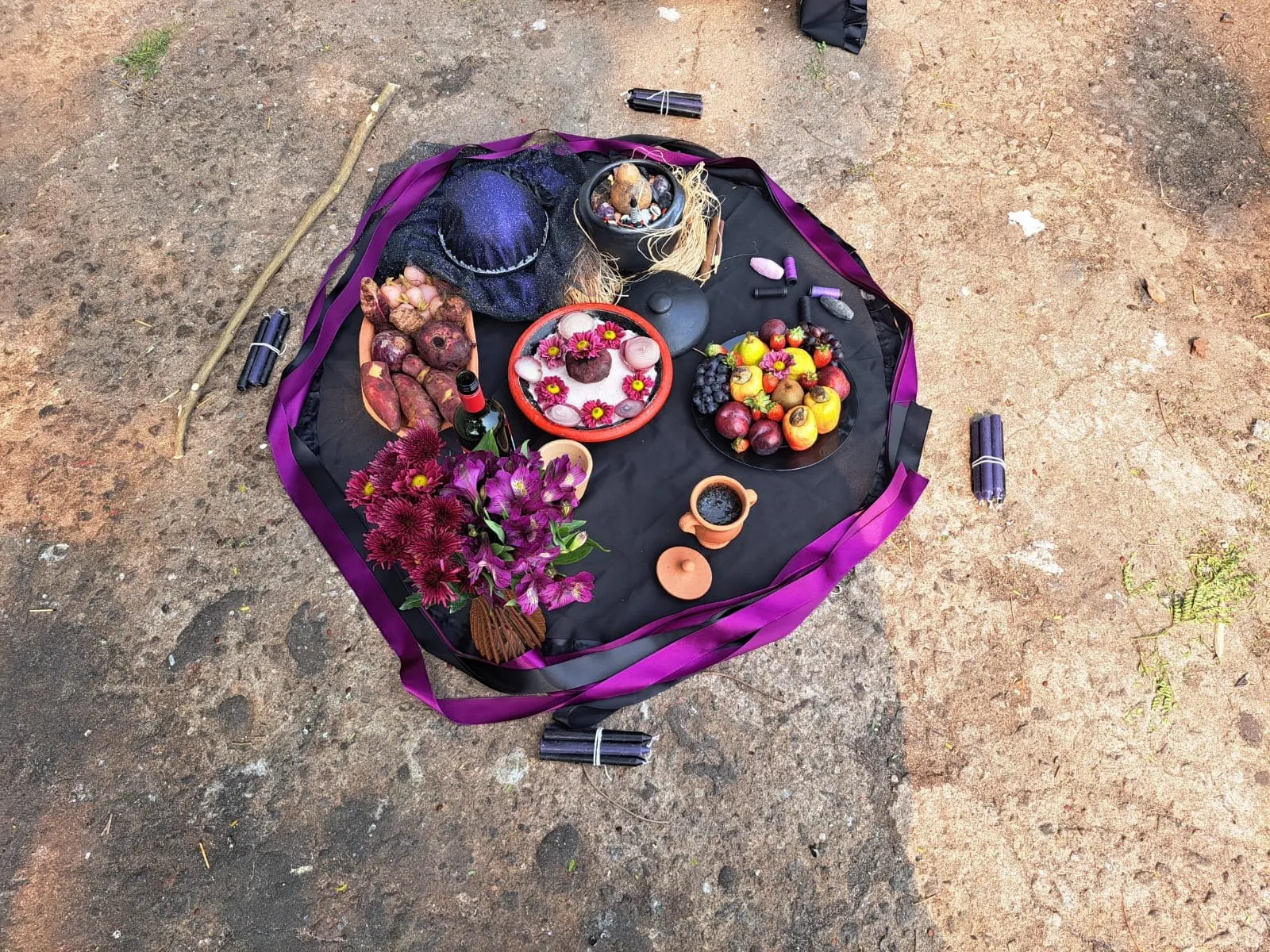 Oferenda a Pombagira vista de cima sobre toalha preta com fita roxa, com flores roxas, chapéu, frutas e velas em volta