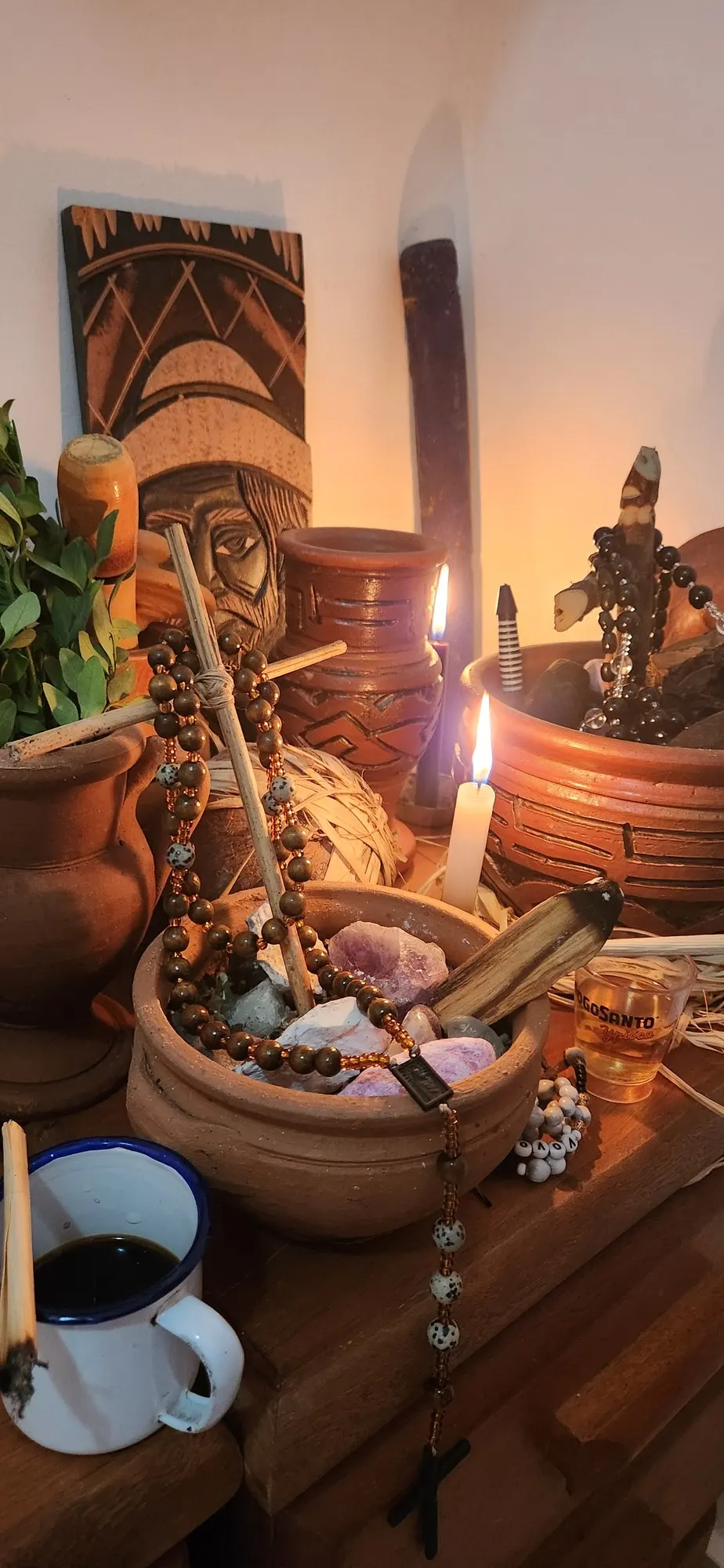 Altar com terço, vela acesa, caneca de café, cristais e potes de barro