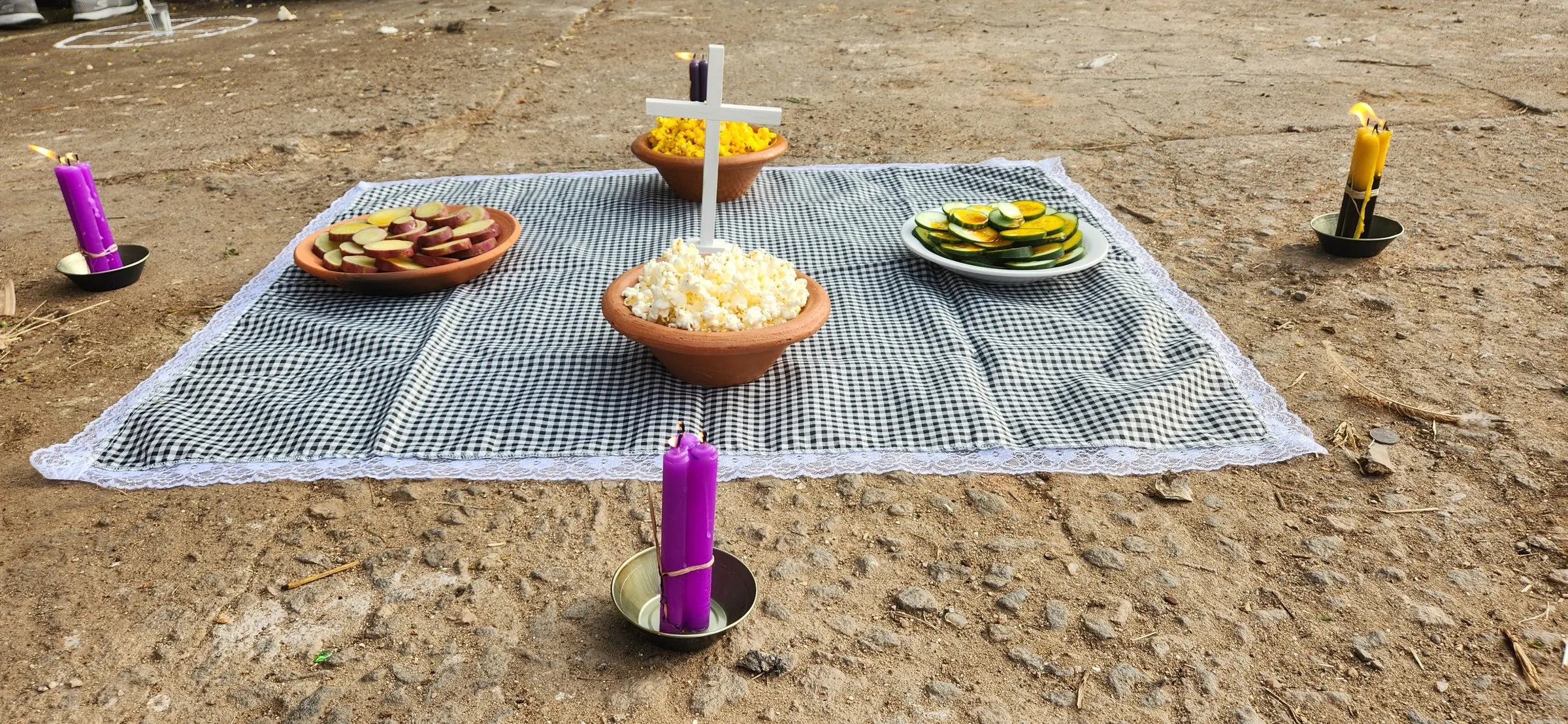 Oferenda na terra: toalha xadrez com rendinha, tigela de pipoca, cruz branca, batata-doce, velas roxas e amarelas
