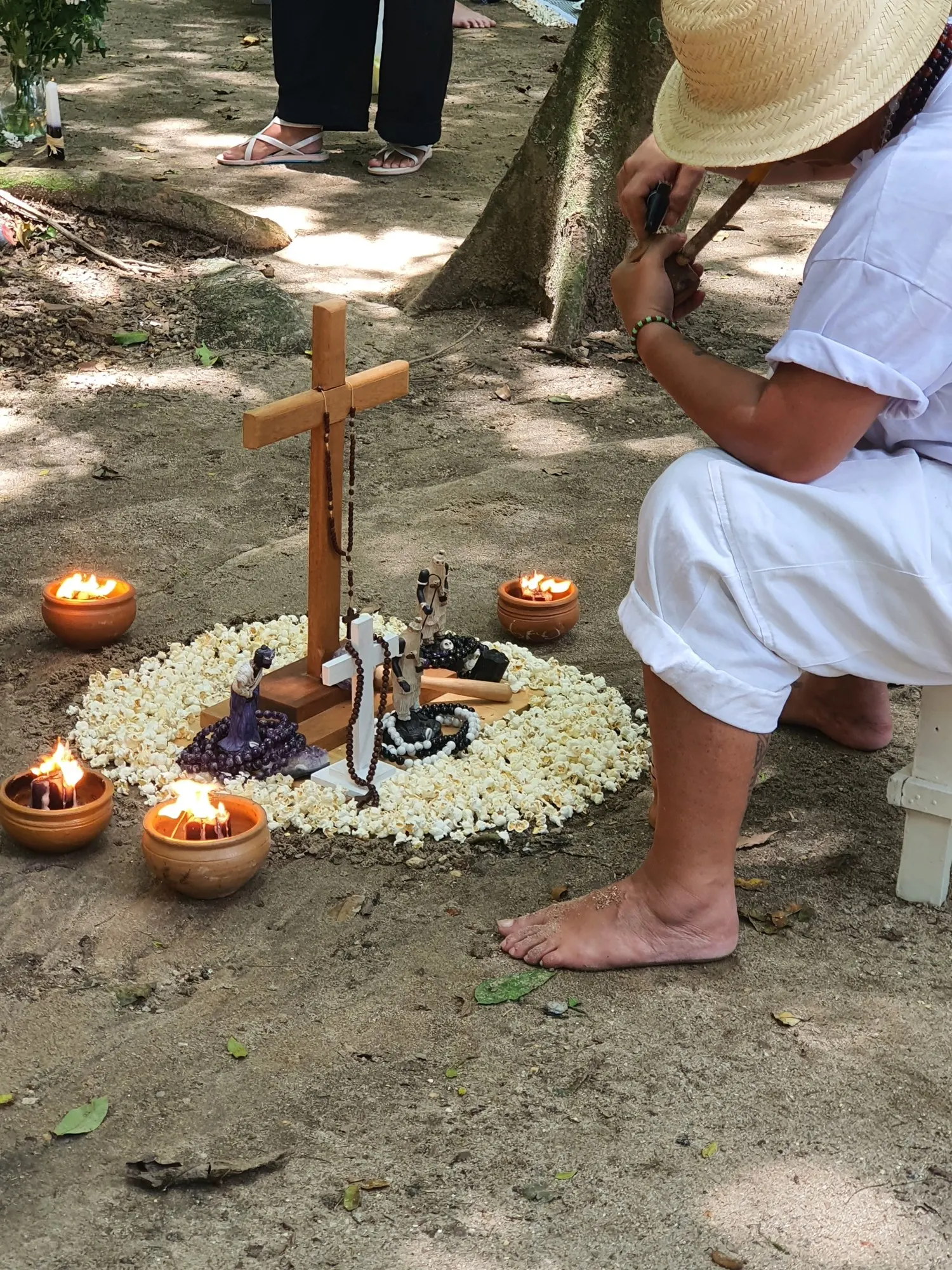 Médium de branco e chapéu de palha, incorporado com Preto Velho, sentado num banquinho, acende o cachimbo diante de uma firmeza com cruz, terço, velas e círculo de pipoca