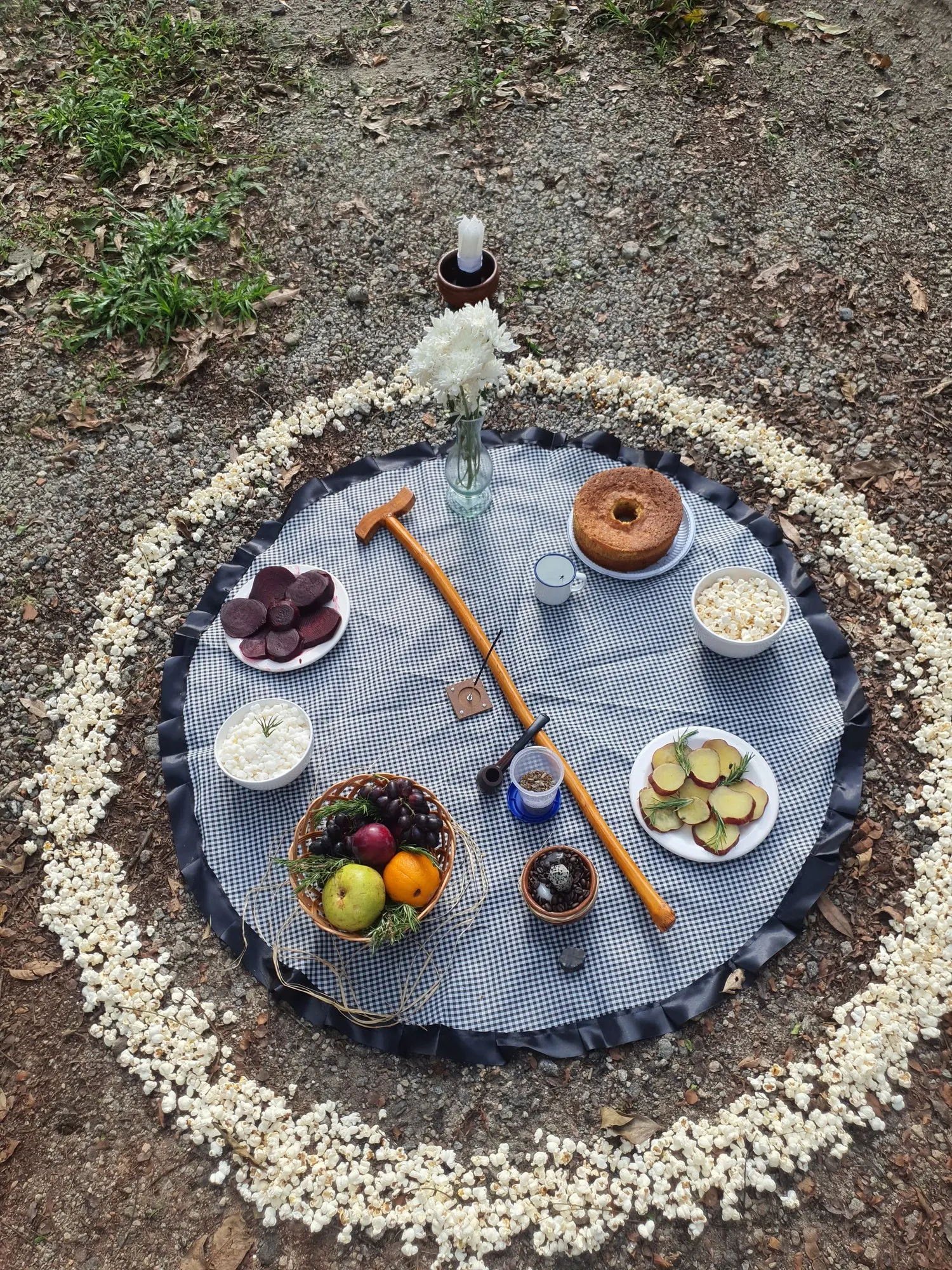 Oferenda vista de cima em toalha xadrez, com bengala de madeira, cesta de frutas e bolo, cercada por um círculo de pipoca