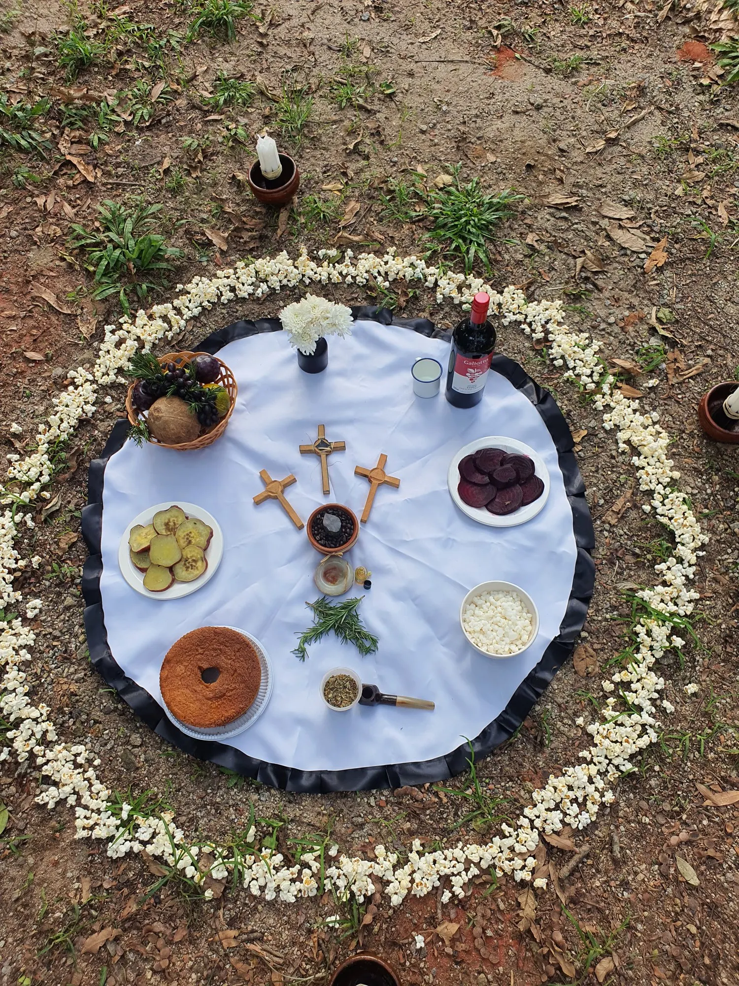 Oferenda vista de cima em toalha branca, com cruzes de madeira, bolo, beterraba e pepino, cercada por um círculo de pipoca
