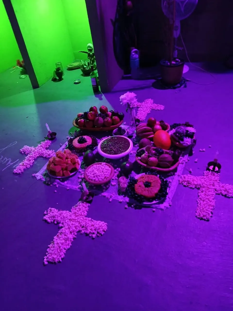 Cruzes de pipoca no chão do templo, sob luz roxa, em volta de pratos com frutas e doces
