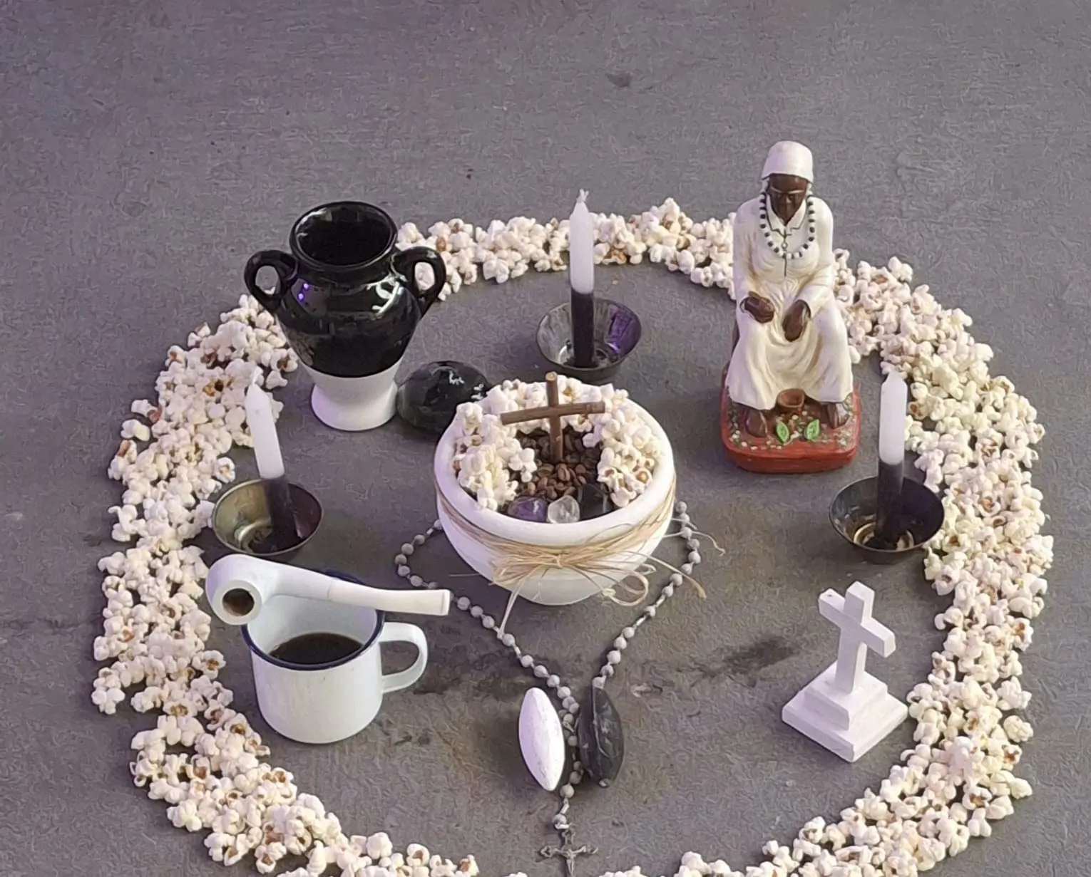 Imagem de Preta Velha, caneca de café, terço, velas e uma cruz branca, dentro de um círculo de pipoca