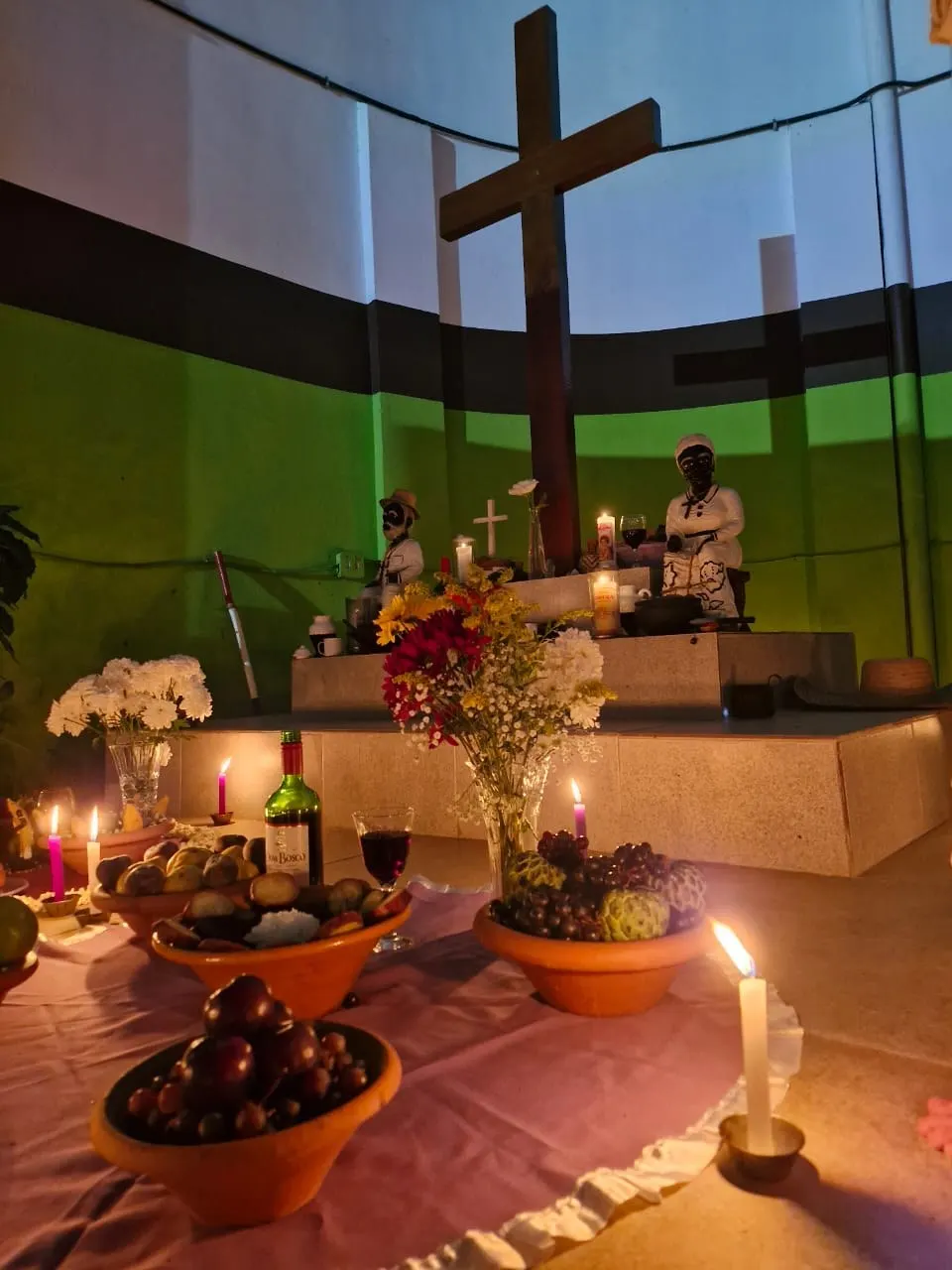 Cruzeiro das Almas do templo com imagens de Pretos Velhos, flores, frutas, vinho e velas acesas sobre toalha lilás