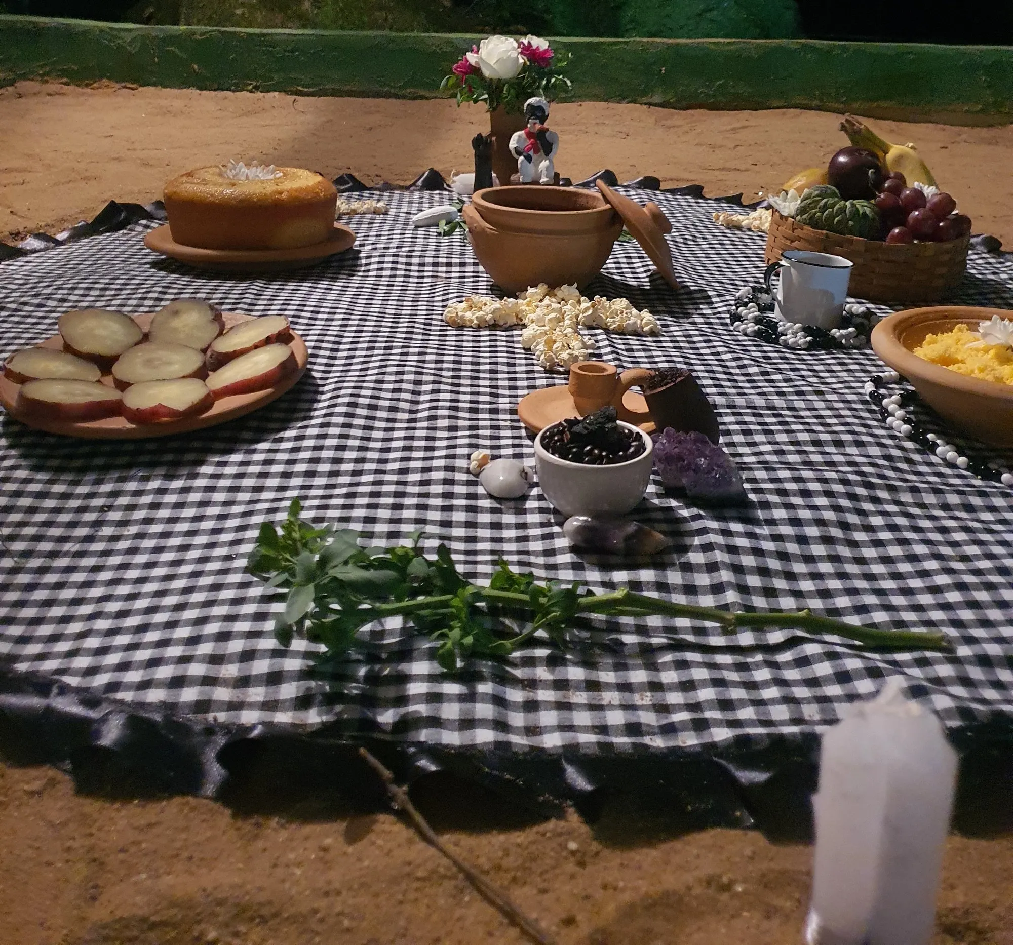 Oferenda à noite sobre toalha xadrez preta e branca, na terra, com bolo, pipoca, frutas, caneca, cristal roxo, flores e uma pequena imagem