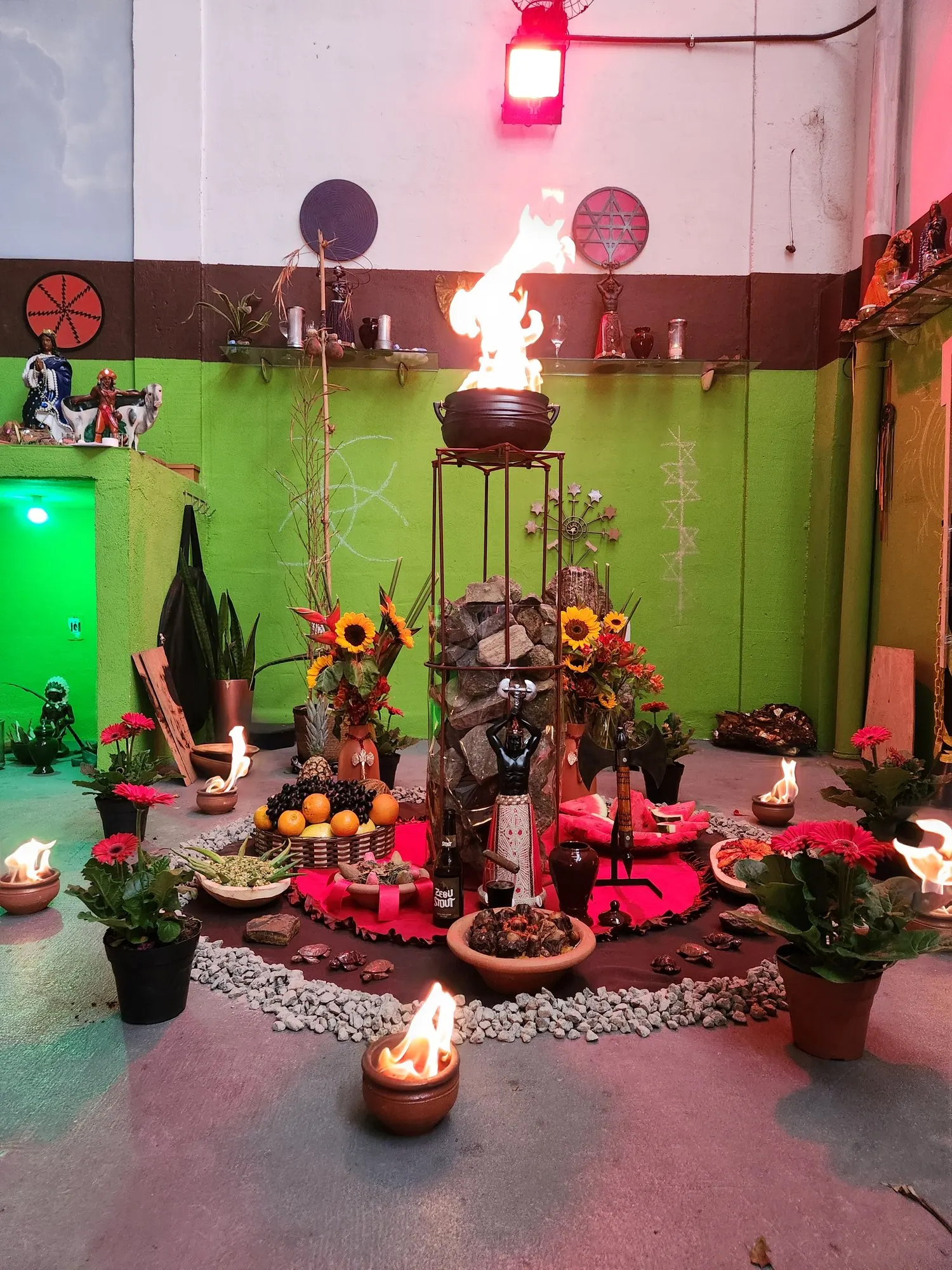 Fogo aceso sobre pedras empilhadas num suporte de ferro, no templo do TEUSP, cercado de girassóis, flores vermelhas e velas