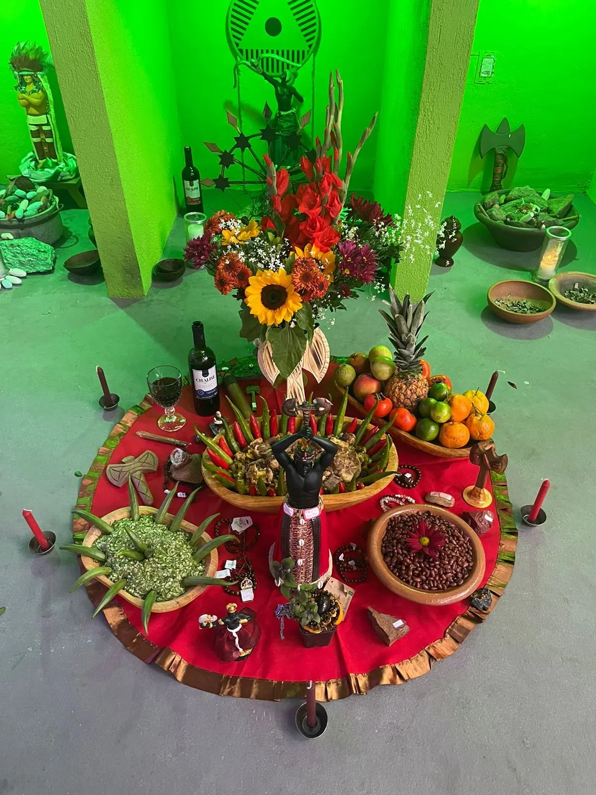 Oferenda a Xangô sobre toalha vermelha no templo, com uma imagem ao centro, girassóis e flores vermelhas, frutas e gamelas com quiabo picado e grãos