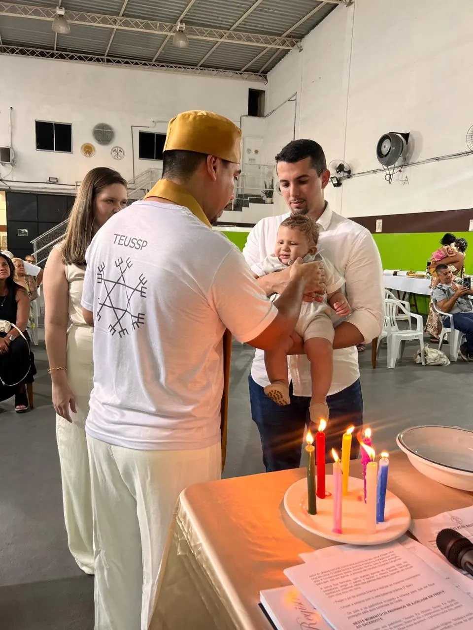 Batismo no templo: o sacerdote abençoa um bebê no colo do pai, com a mãe ao lado e velas coloridas acesas na mesa