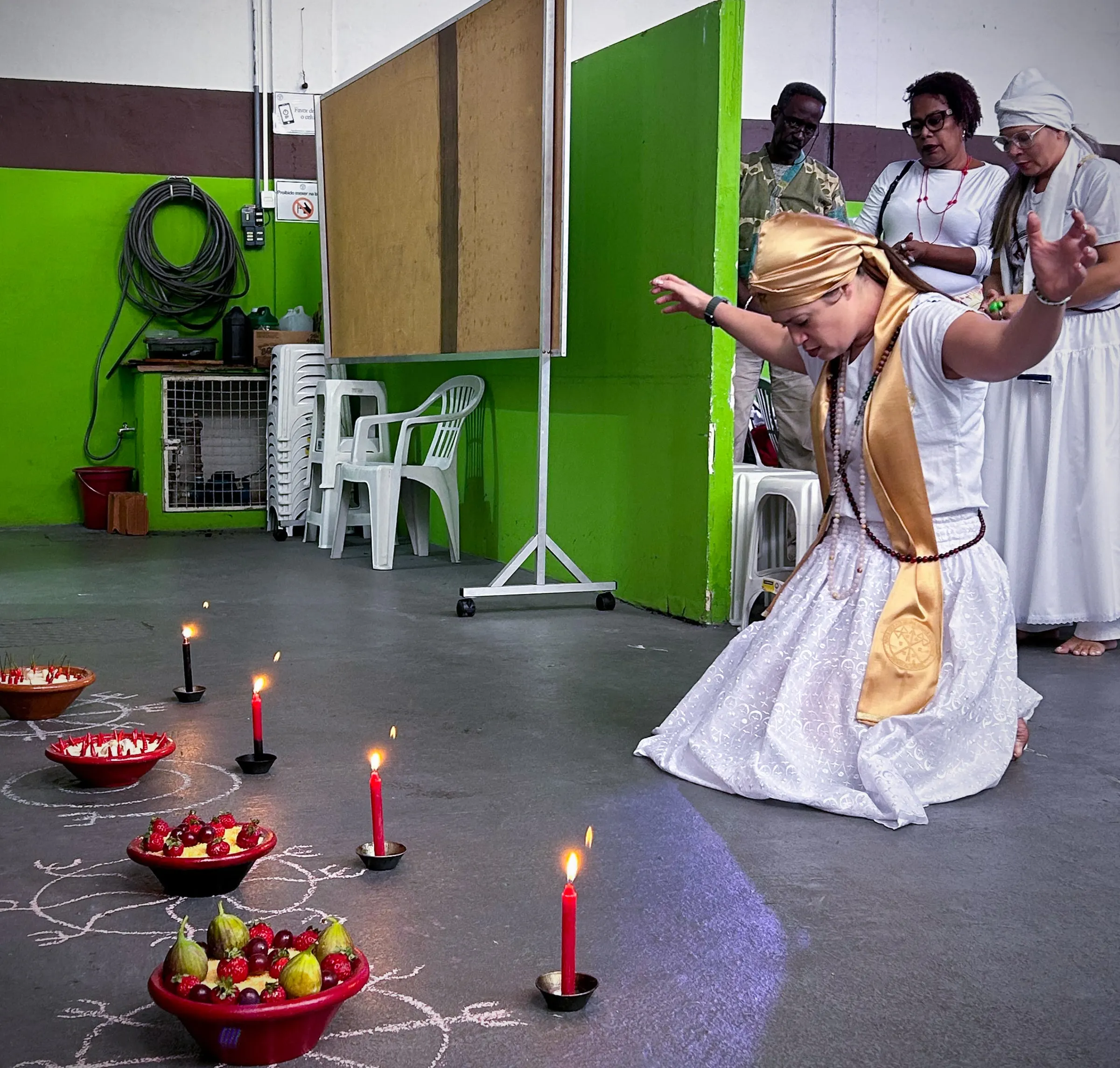 Formanda ajoelhada, de estola dourada e braços abertos, diante de oferendas de frutas e velas vermelhas no chão do templo
