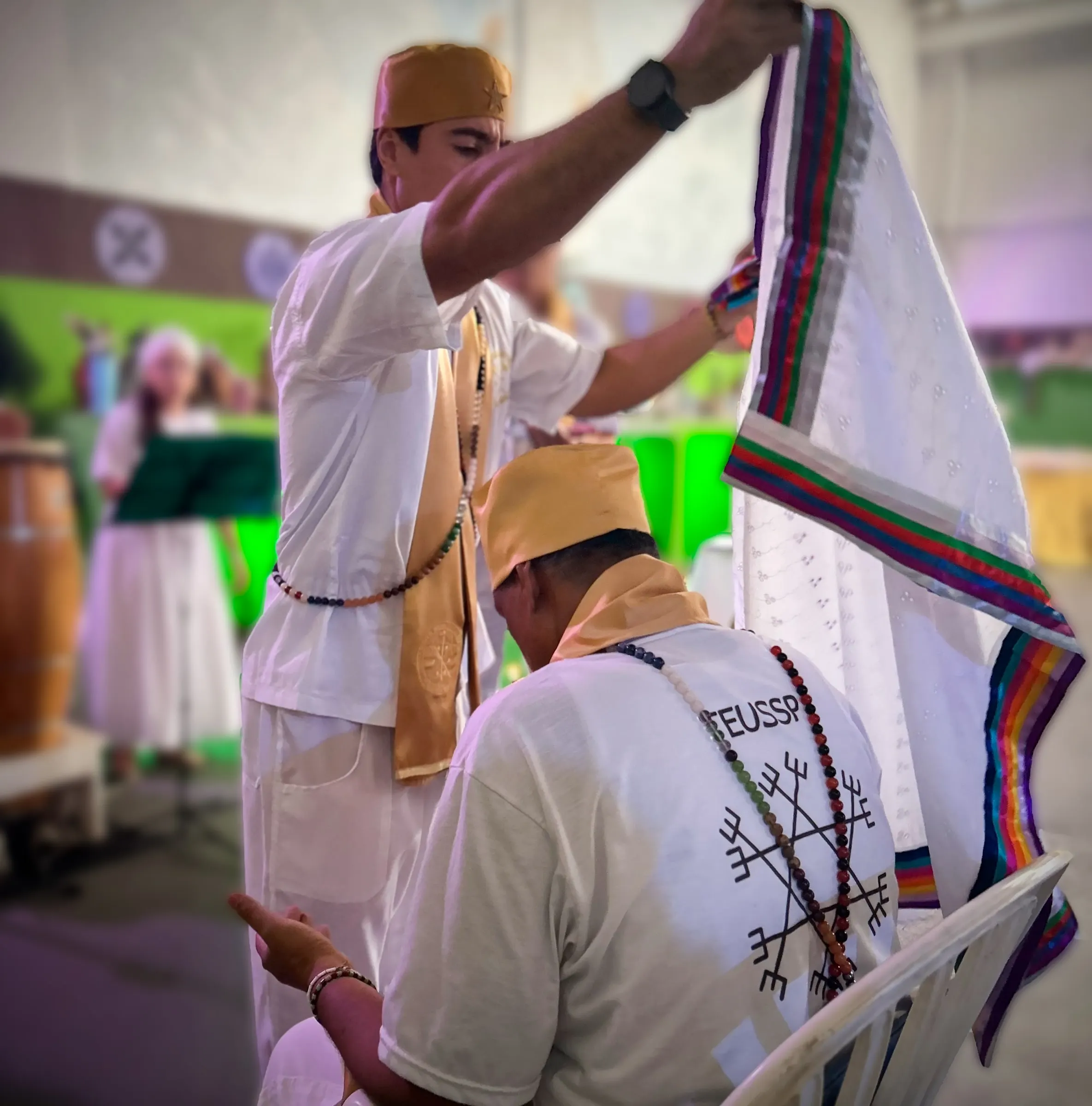 Um sacerdote estende um pano branco de barra colorida sobre um formando sentado, de turbante e estola dourados