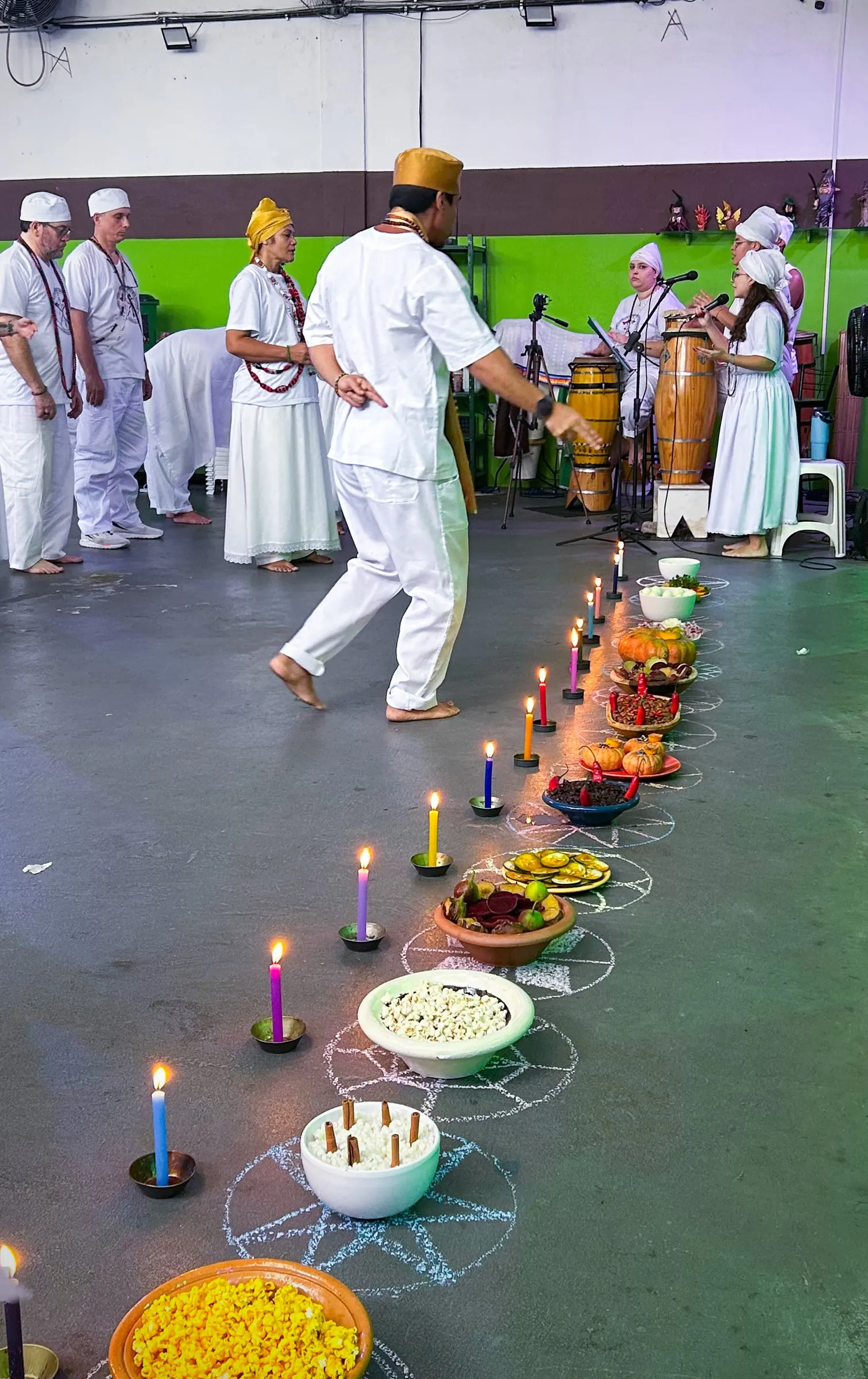 Na formatura, um sacerdote dança ao lado de um caminho de velas acesas e oferendas no chão, com os médiuns e os atabaques ao fundo