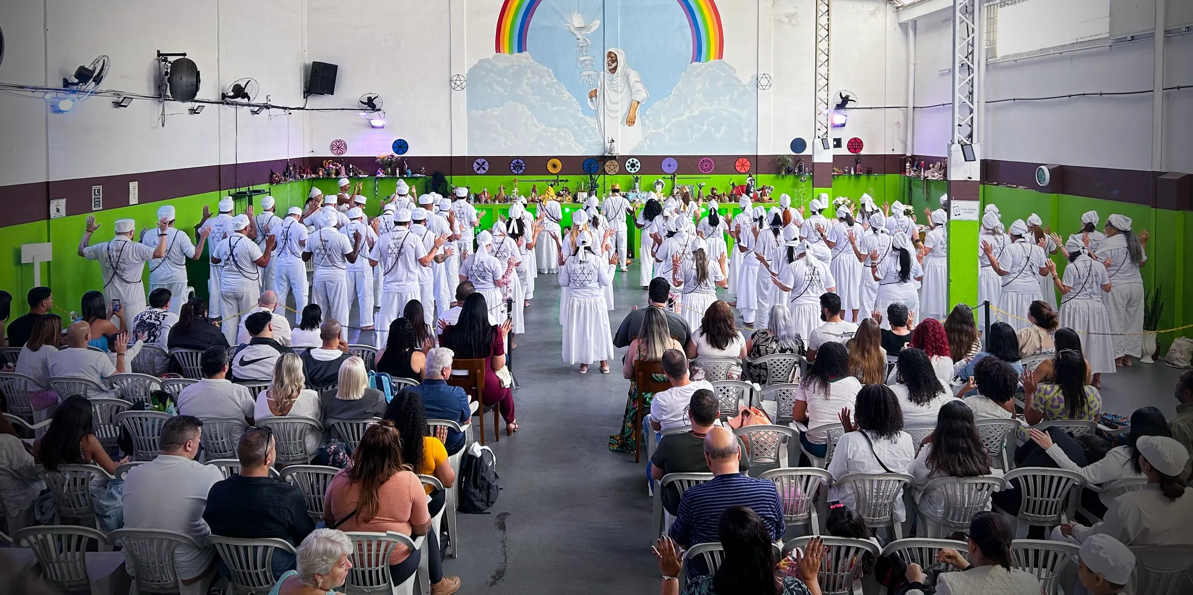 Templo cheio na formatura da primeira turma da formação sacerdotal, em 6 de julho de 2025: médiuns de branco diante do mural do arco-íris e o público sentado