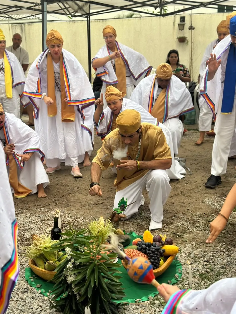 Na formatura da Turma 2, Bruno Amorim, de dourado, ajoelhado diante das oferendas de frutas, espigas de milho e folhas no chão, sopra fumaça sobre elas, cercado pelos formandos de branco com estolas coloridas e turbantes dourados