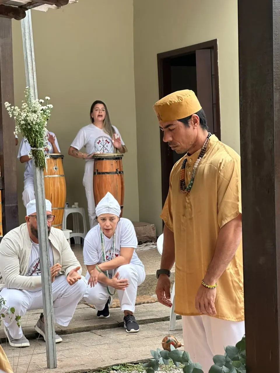 Bruno Amorim, de dourado, conduz a gira de formatura; atrás dele, a curimba toca os atabaques e dois médiuns de branco acompanham agachados