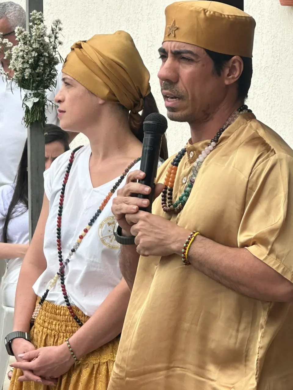 Flávia Amorim, de turbante dourado, ao lado de Bruno Amorim, de dourado, que fala ao microfone na formatura da Turma 2