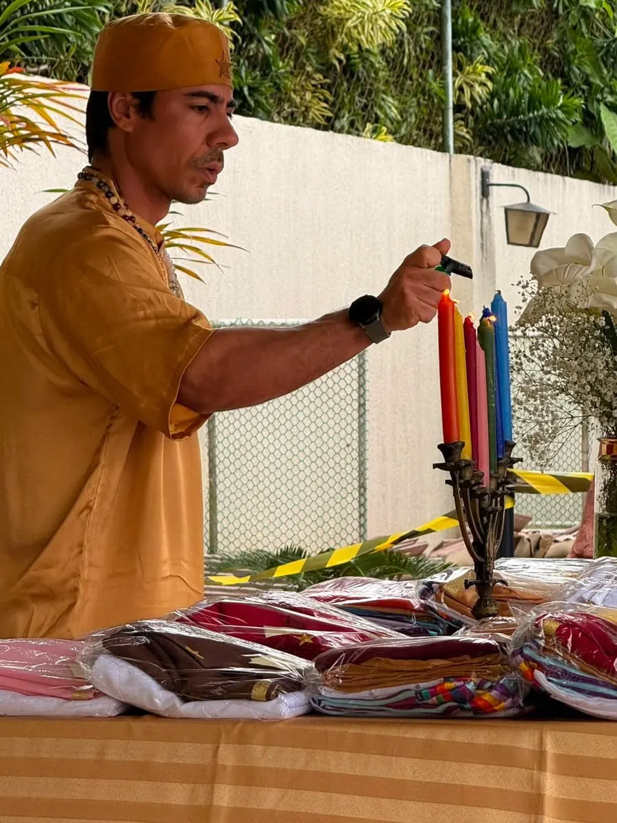 Bruno Amorim acende velas coloridas num castiçal, sobre a mesa com as roupas e estolas dobradas para a formatura