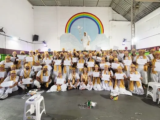 Turma da formação sacerdotal reunida, com roupas brancas, estolas e turbantes dourados, diante de um mural de arco-íris