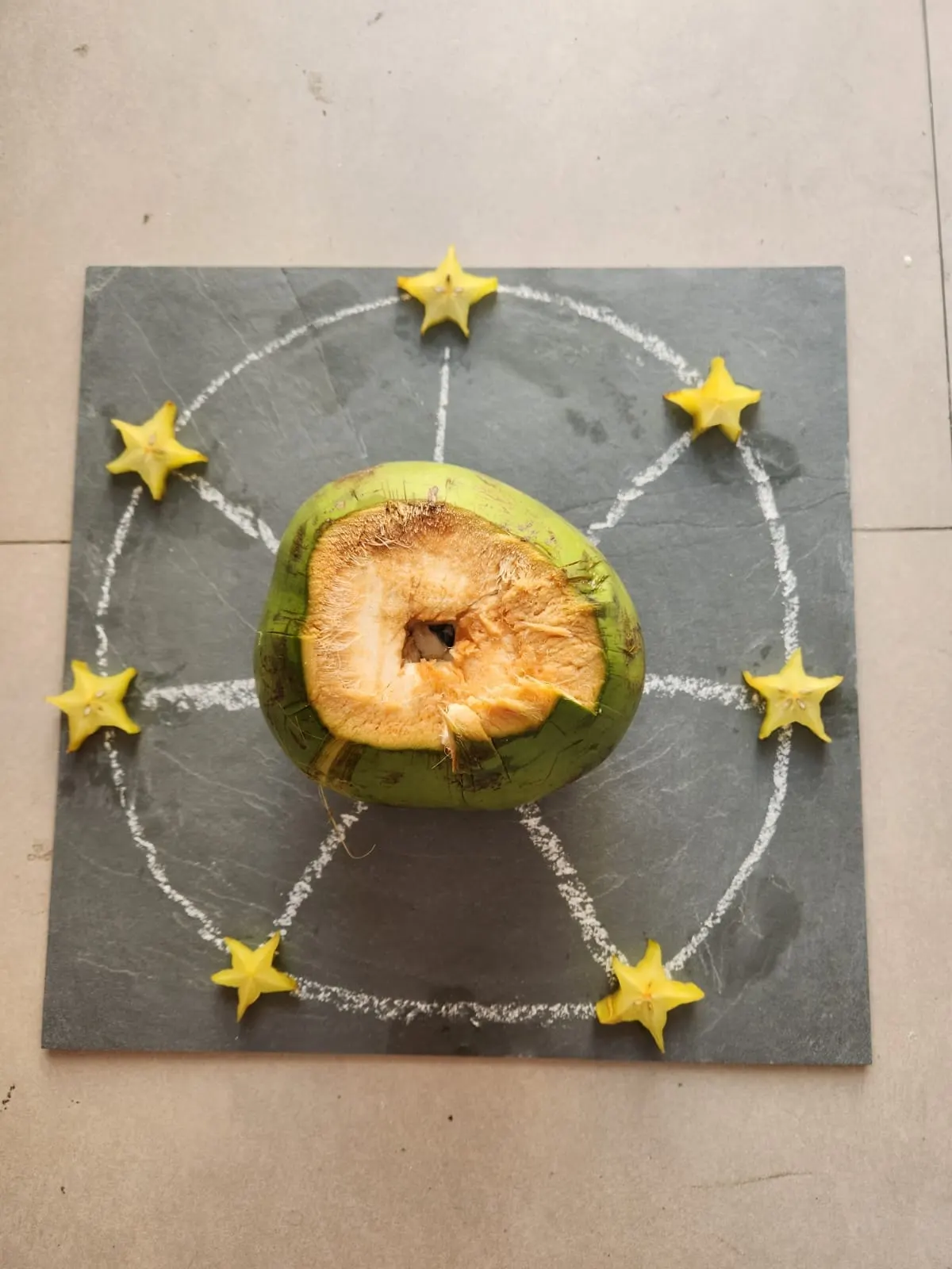 Círculo riscado a giz sobre uma placa de ardósia, com um coco verde aberto no centro e sete fatias de carambola ao redor