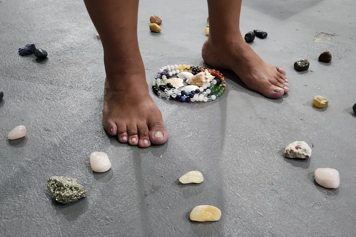 Pés descalços no meio de pedras e cristais espalhados pelo chão, com uma pequena mandala de contas coloridas e conchas entre os pés