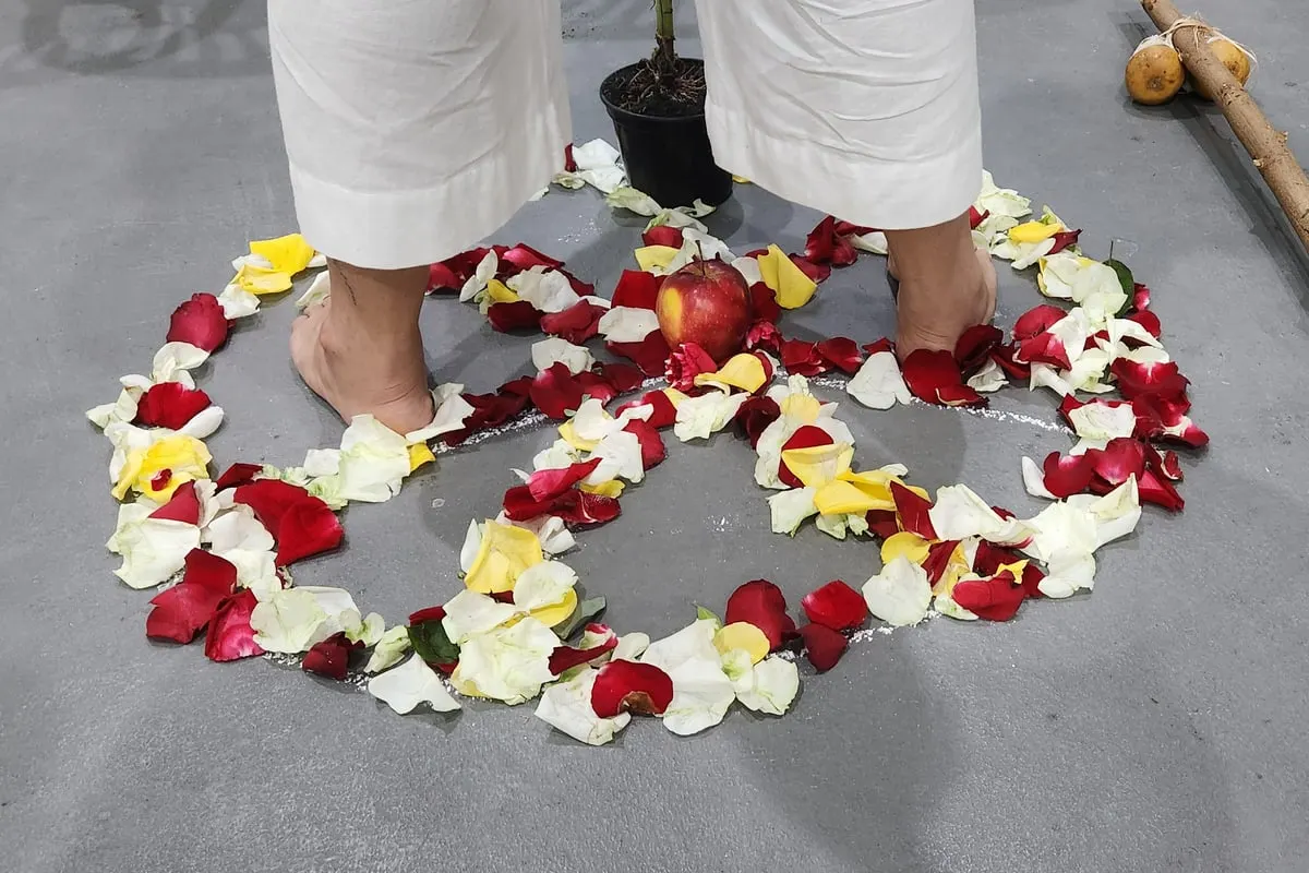 Pés descalços de uma pessoa de roupa branca dentro de um círculo de pétalas de rosas vermelhas, brancas e amarelas, com uma maçã no centro