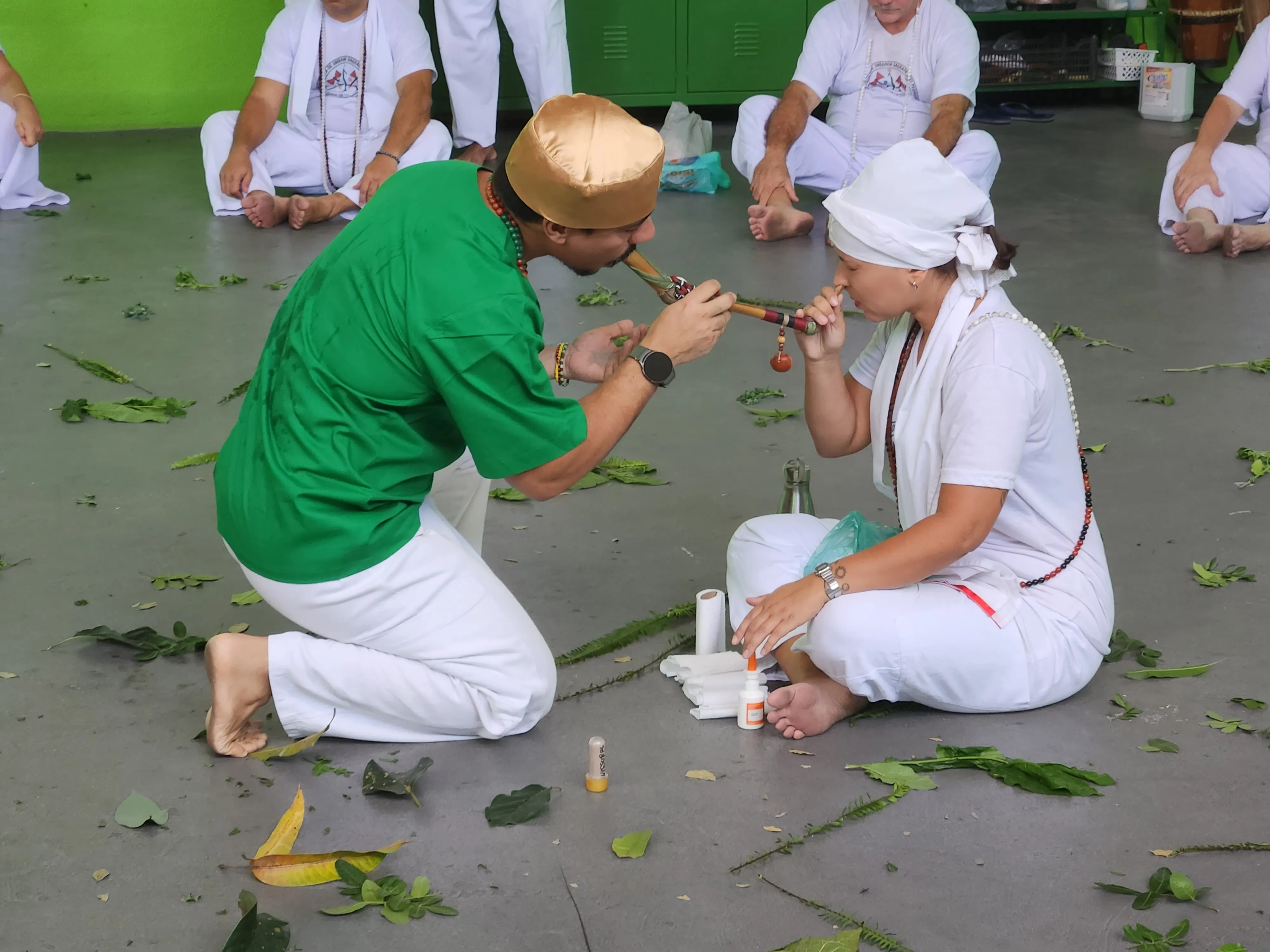 Roda de rapé no TEUSP: ajoelhado, um homem de camisa verde e gorro dourado aplica rapé, com um aplicador longo, numa participante vestida de branco; ao fundo, outras pessoas de branco sentadas no chão, entre folhas espalhadas.