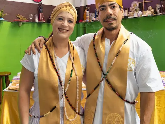 Flávia Amorim e Bruno Amorim, sorridentes, com estolas douradas e fios de contas, diante do congá