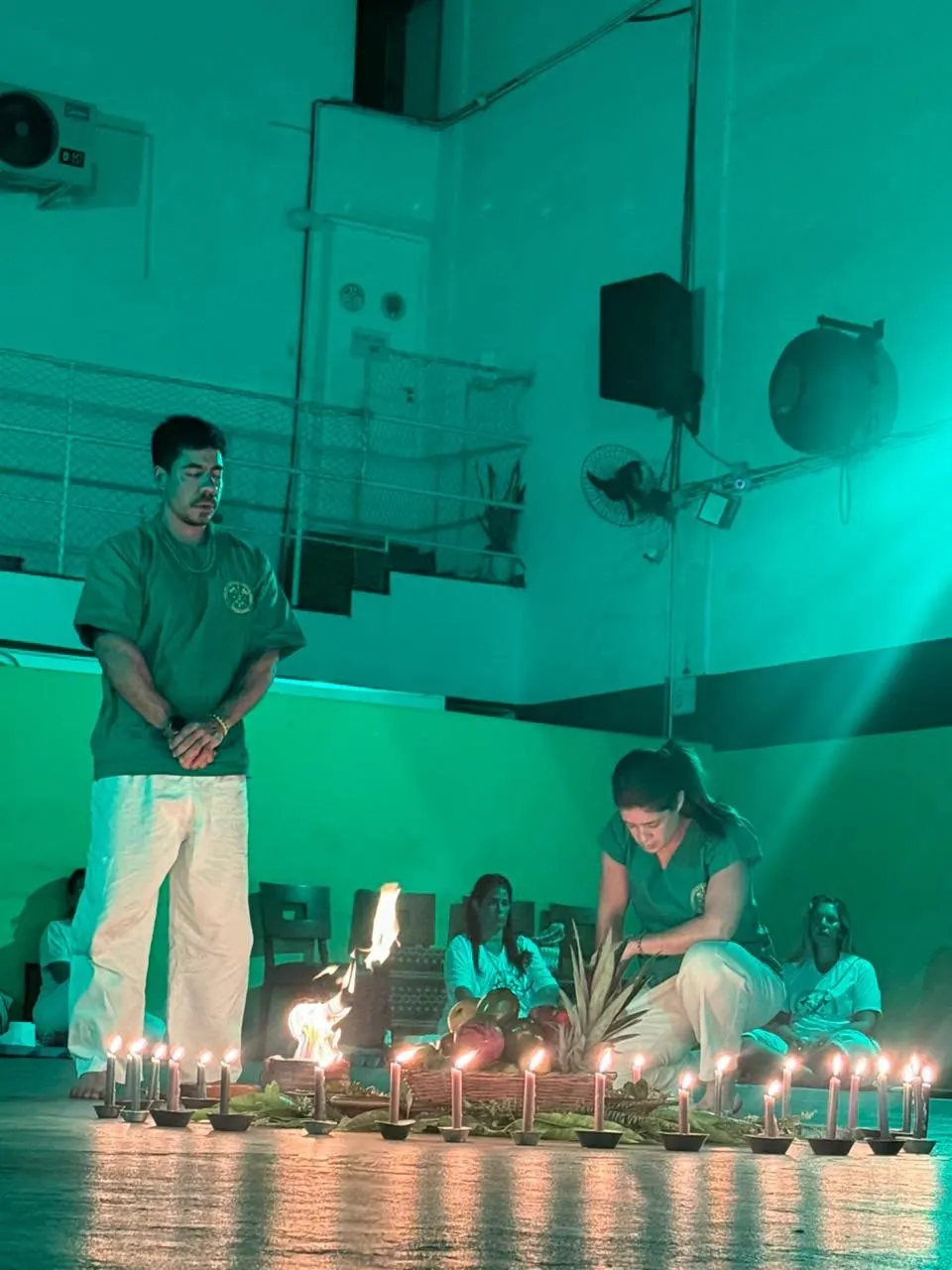 Roda de rapé sob luz verde: Pai Bruno Amorim, de pé e de olhos fechados, ao lado de uma fileira de velas acesas e de uma pequena fogueira; uma mulher ajoelhada arruma as frutas, e os participantes, de branco, estão sentados ao fundo