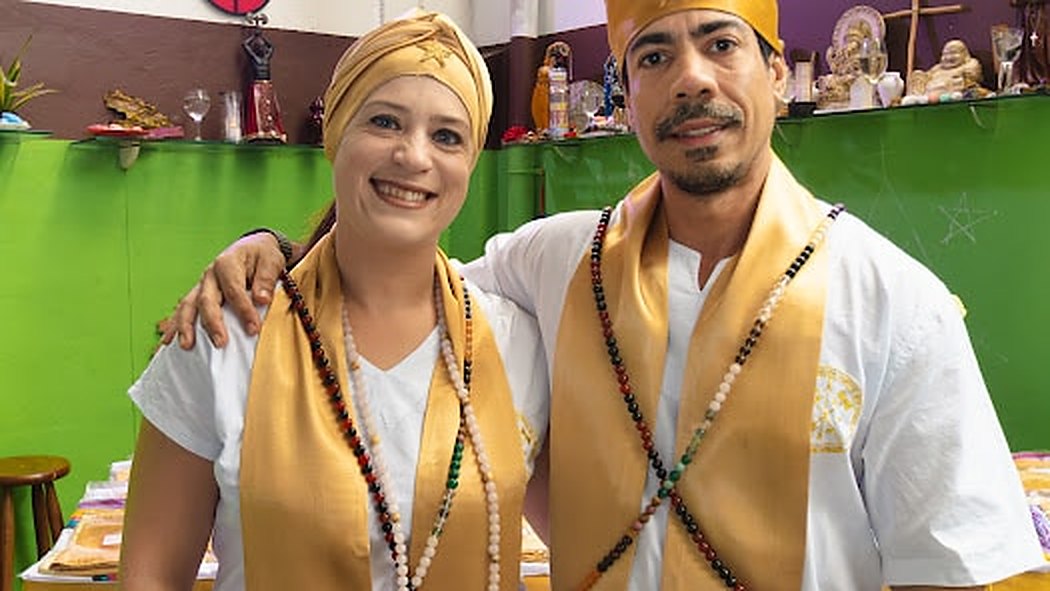 Flávia Amorim e Bruno Amorim, sorridentes, com estolas douradas e fios de contas, diante do congá