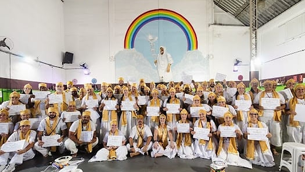 Turma da formação sacerdotal reunida, com roupas brancas, estolas e turbantes dourados, diante de um mural de arco-íris