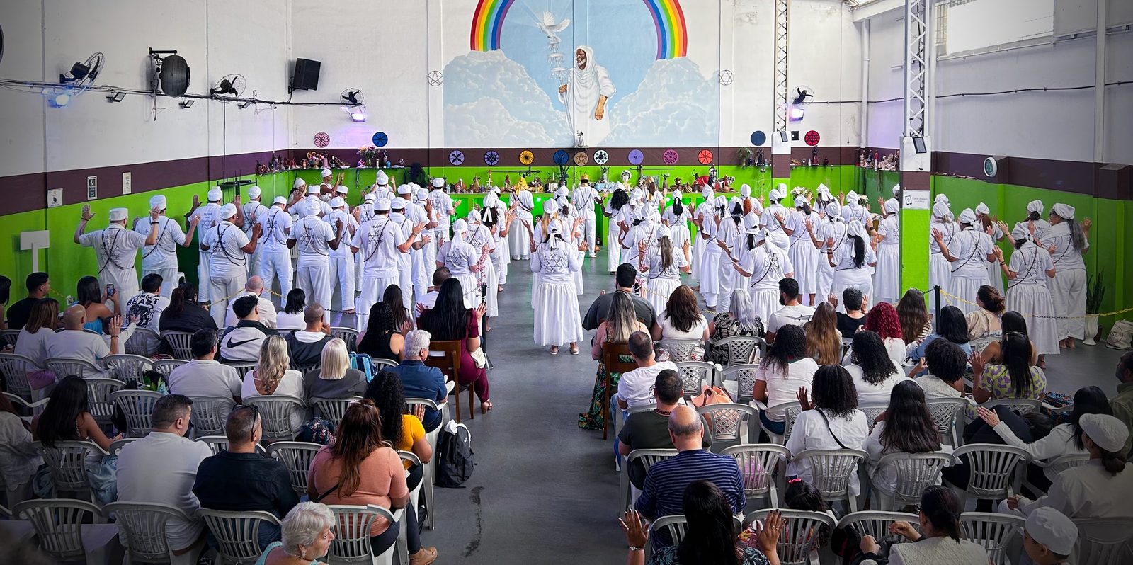 Templo do TEUSP cheio na formatura sacerdotal da Turma 1: médiuns de branco diante do mural de arco-íris e o público sentado